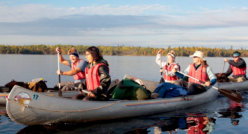 A group of students wearing life jackets smile as they paddle canoes on calm water. In the distance, trees line the shore. 